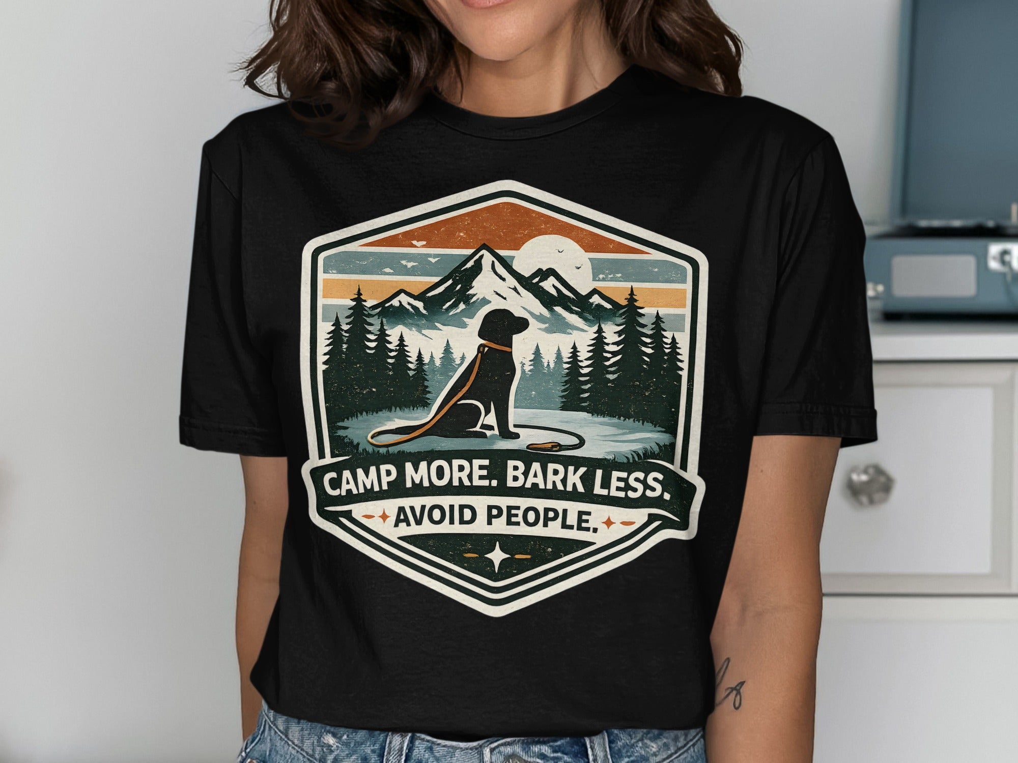 Black t-shirt with a badge design featuring a dog, mountains, and the text "Camp More, Bark Less, Avoid People."